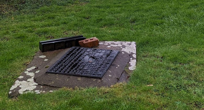 A damp grassy field in South Wales features a manhole in the foreground with a red brick building after rainfall